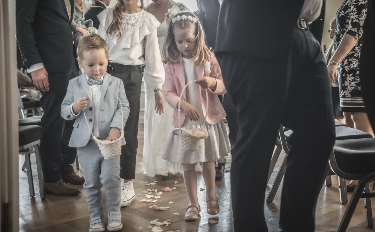 Kinder in festlicher Kleidung streuen Blumenblätter auf einem Gang, umgeben von Erwachsenen.