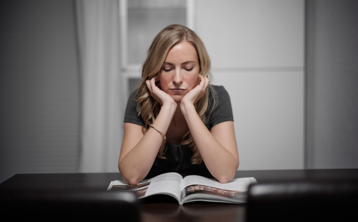 Frau sitzt an einem Tisch und liest ein Buch, stützt den Kopf auf ihre Hände.