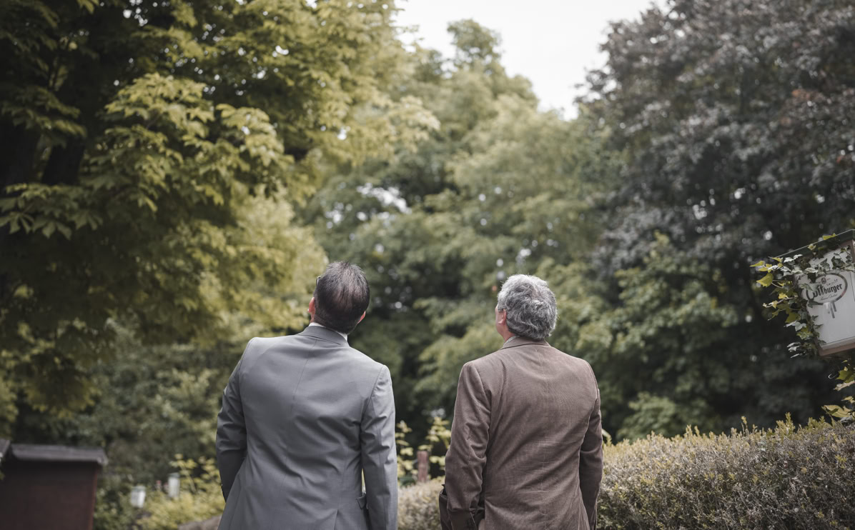 Zwei Personen in Anzügen stehen im Freien und schauen in Richtung der Bäume.