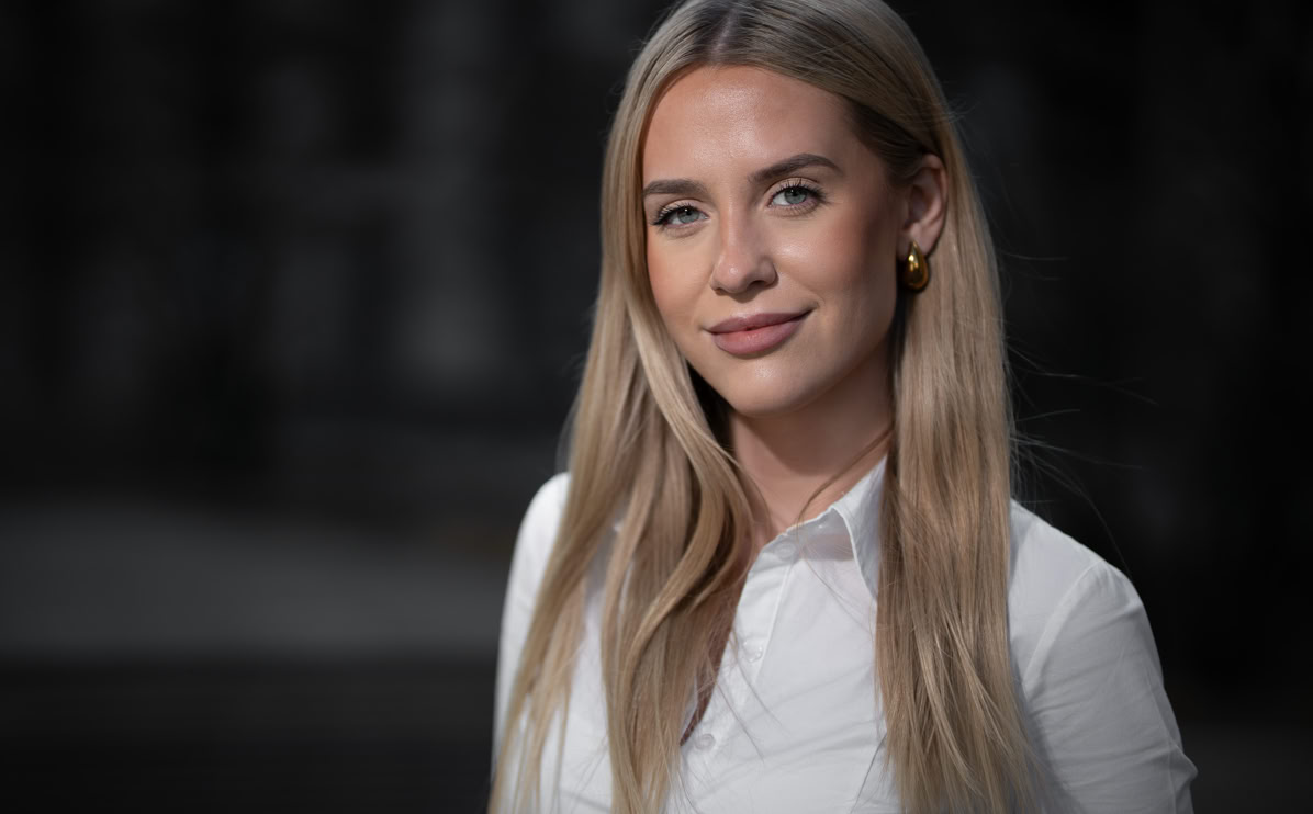 Eine Frau mit langen blonden Haaren trägt ein weißes Hemd und steht vor einem unscharfen Hintergrund.