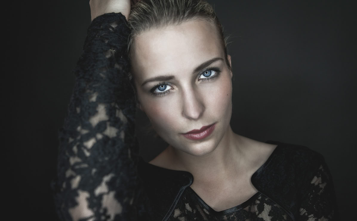 Frau mit blonden Haaren und blauer Spitze, die in die Kamera schaut, vor dunklem Hintergrund.