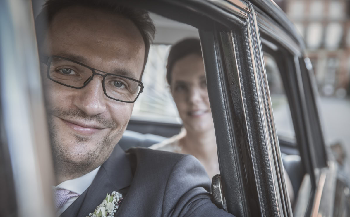 Mann mit Brille und Anzug sitzt in einem Auto, eine Frau ist im Hintergrund zu sehen.
