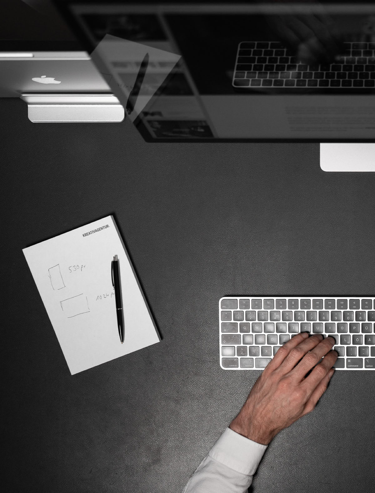Zwei Hände auf einer Computertastatur, ein Monitor, ein Notizblock mit Stift und ein Computer auf einem dunklen Schreibtisch.
