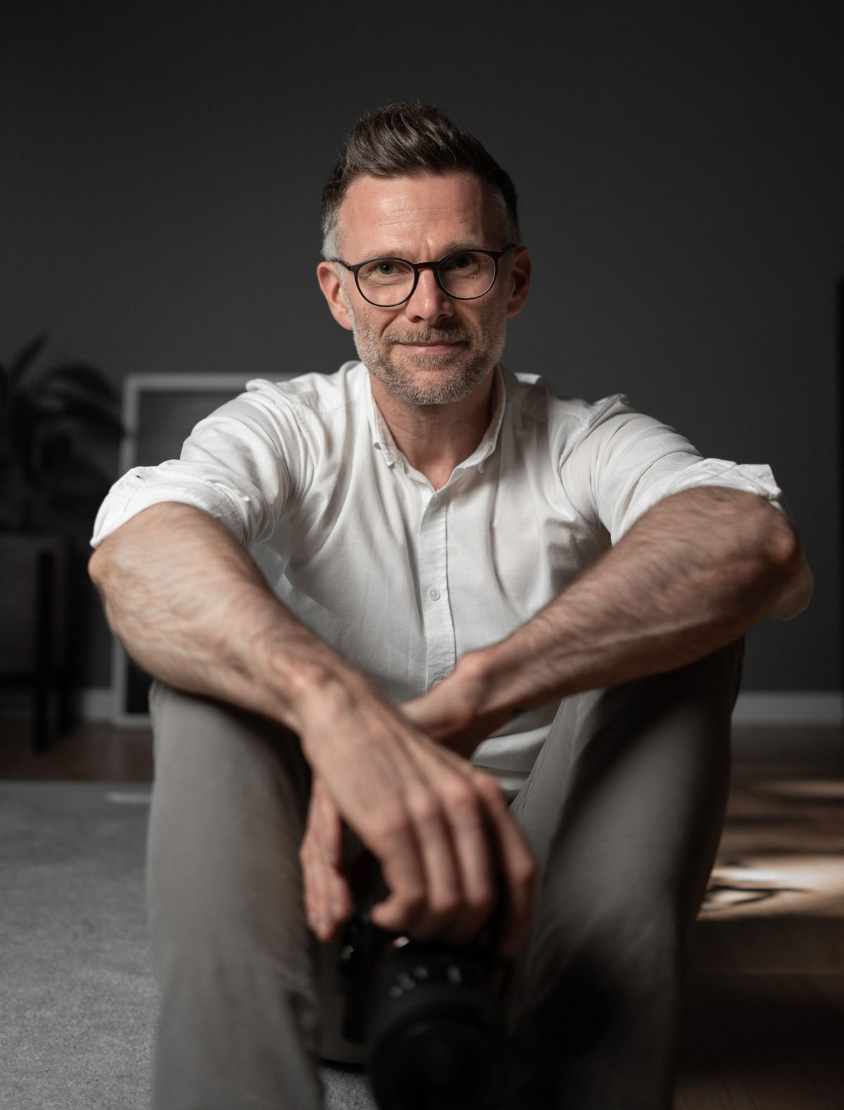 Ein Mann mit Brille und weißem Hemd sitzt auf dem Boden und hält eine Kamera.