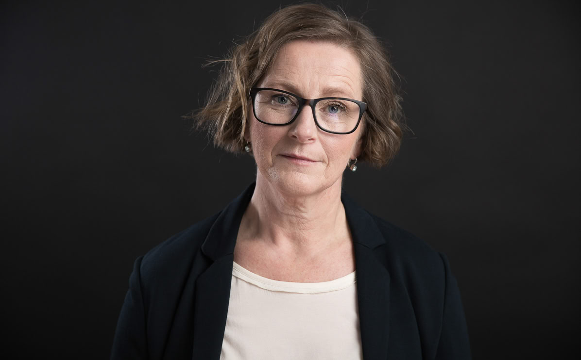 Eine Frau mit Brille und kurzen braunen Haaren trägt ein weißes Oberteil und einen dunklen Blazer vor einem schwarzen Hintergrund.