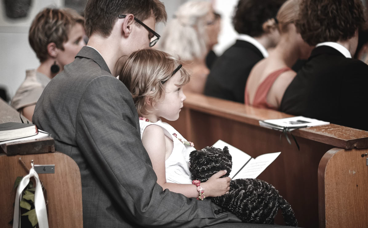 Ein Mann in einem Anzug sitzt auf einer Kirchenbank und hält ein kleines Mädchen auf dem Schoß, das ein Buch in der Hand hält. Im Hintergrund sind weitere Personen auf Bänken zu sehen.