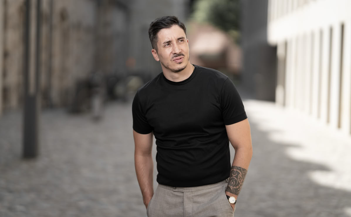 Ein Mann in einem schwarzen T-Shirt und beigen Hosen steht auf einer gepflasterten Straße und schaut zur Seite.
