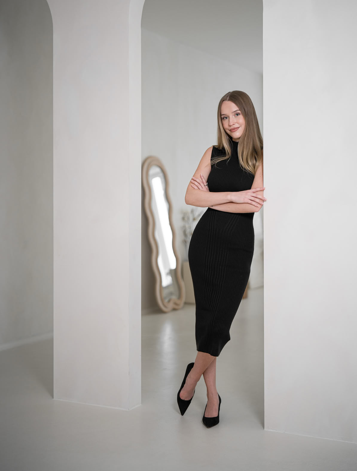 Eine Frau in einem schwarzen Kleid lehnt an einem weißen Bogen in einem minimalistischen Raum mit einem großen Spiegel im Hintergrund.