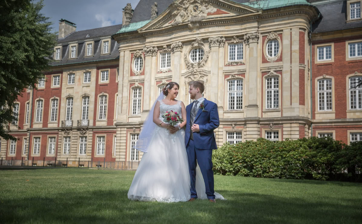 Ein Brautpaar steht auf einer Wiese vor einem historischen Gebäude. Die Braut trägt ein weißes Kleid und hält einen Blumenstrauß, der Bräutigam trägt einen blauen Anzug.