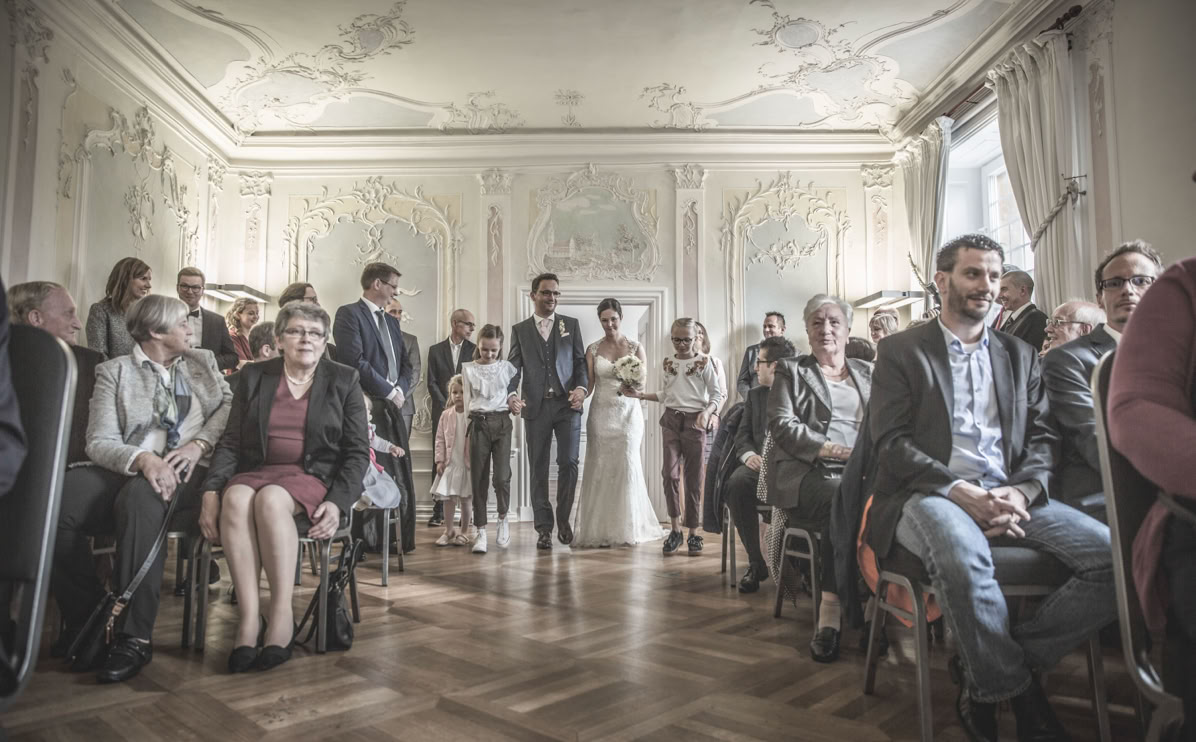 Eine Hochzeitszeremonie in einem eleganten Raum mit dekorativen Wänden, bei der ein Brautpaar den Gang entlang schreitet, umgeben von sitzenden Gästen.