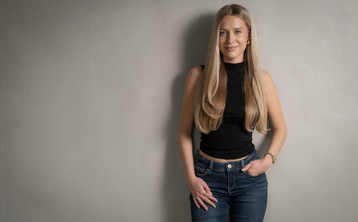 Frau mit langen blonden Haaren, schwarzem ärmellosen Oberteil und blauer Jeans steht vor einer grauen Wand.