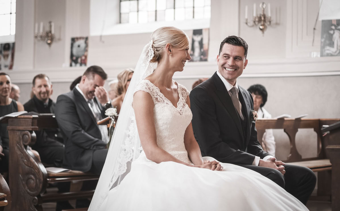 Braut und Bräutigam sitzen lächelnd nebeneinander in einer Kirche, umgeben von Gästen.