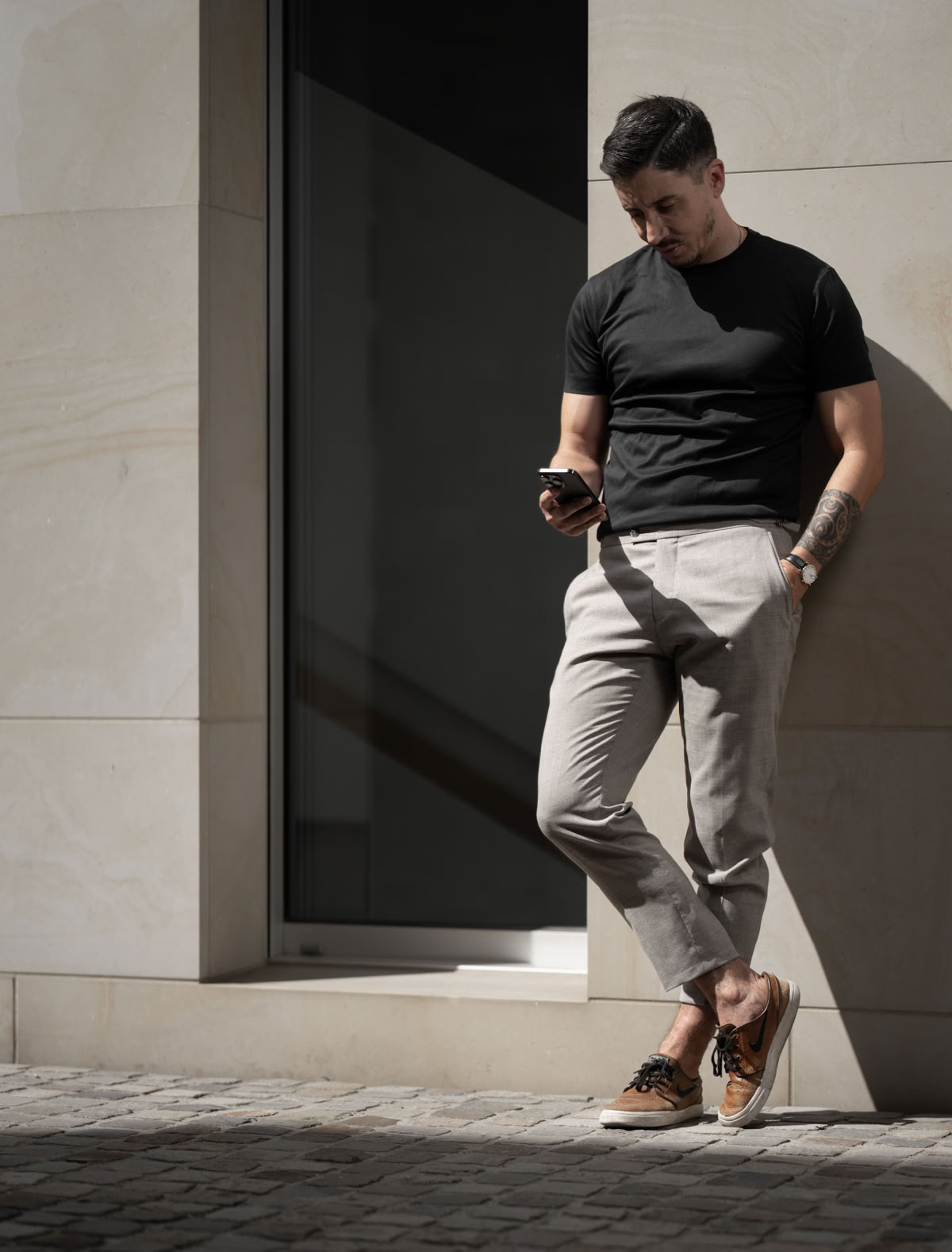 Ein Mann lehnt an einer Wand, trägt ein schwarzes T-Shirt und graue Hosen, schaut auf ein Smartphone in seiner Hand.
