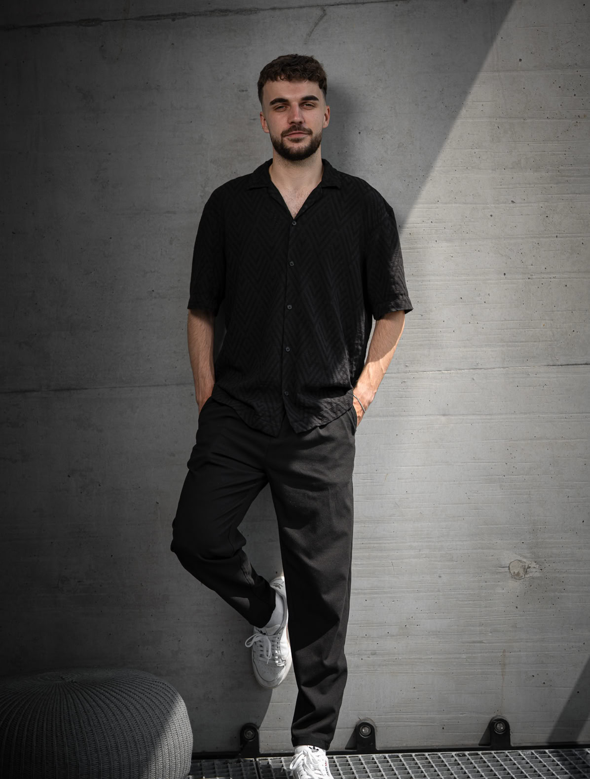 Eine Person in schwarzer Kleidung lehnt an einer grauen Betonwand, mit den Händen in den Hosentaschen, weiße Turnschuhe tragend.