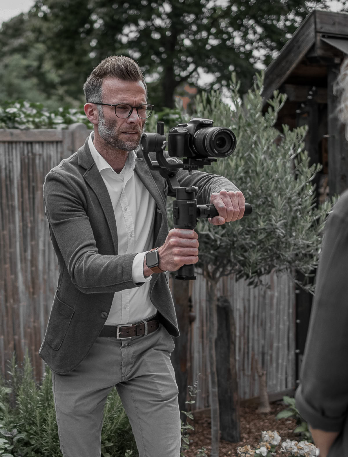 Ein Mann mit Brille und Bart hält eine Kamera auf einem Stabilisator und filmt eine Frau mit lockigem Haar im Freien. Im Hintergrund sind ein Holzzaun und Bäume zu sehen.