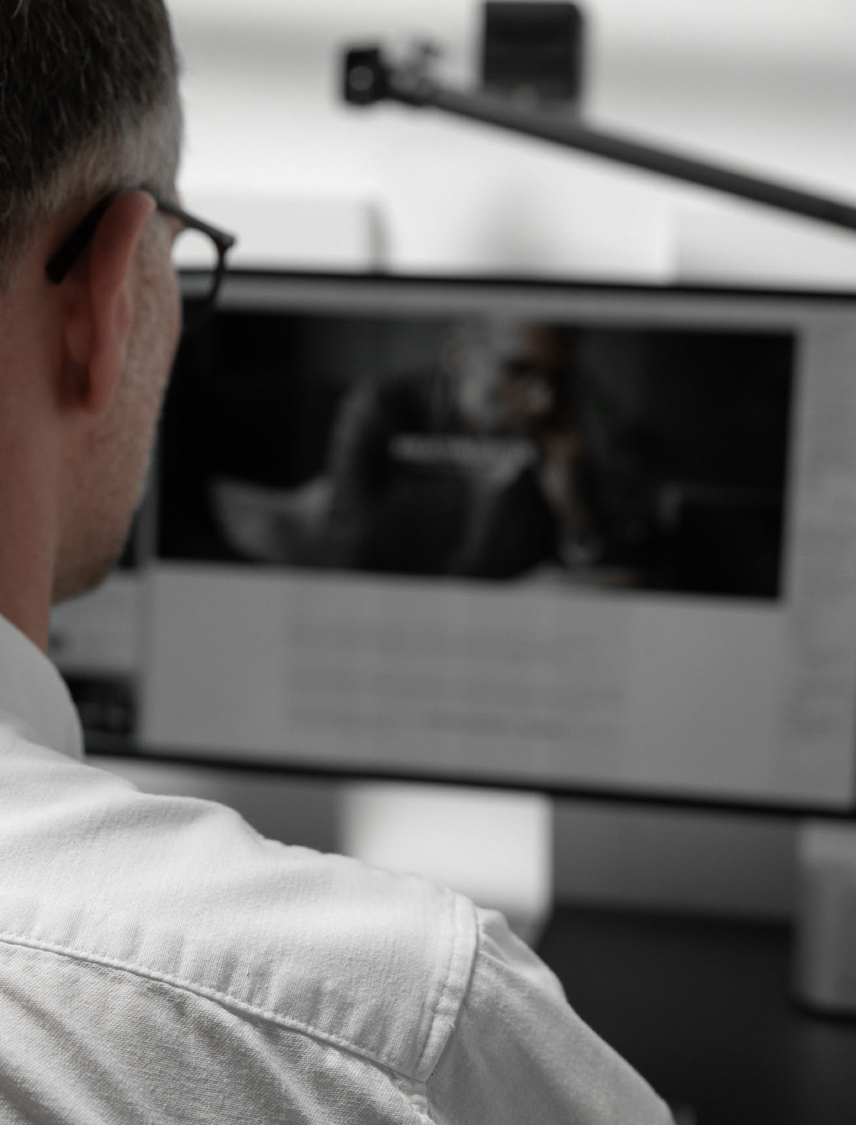 Eine Person in einem weißen Hemd sitzt vor einem Computerbildschirm, auf dem ein Video oder Bild angezeigt wird. Daneben befindet sich eine Kamera auf einem Stativ.