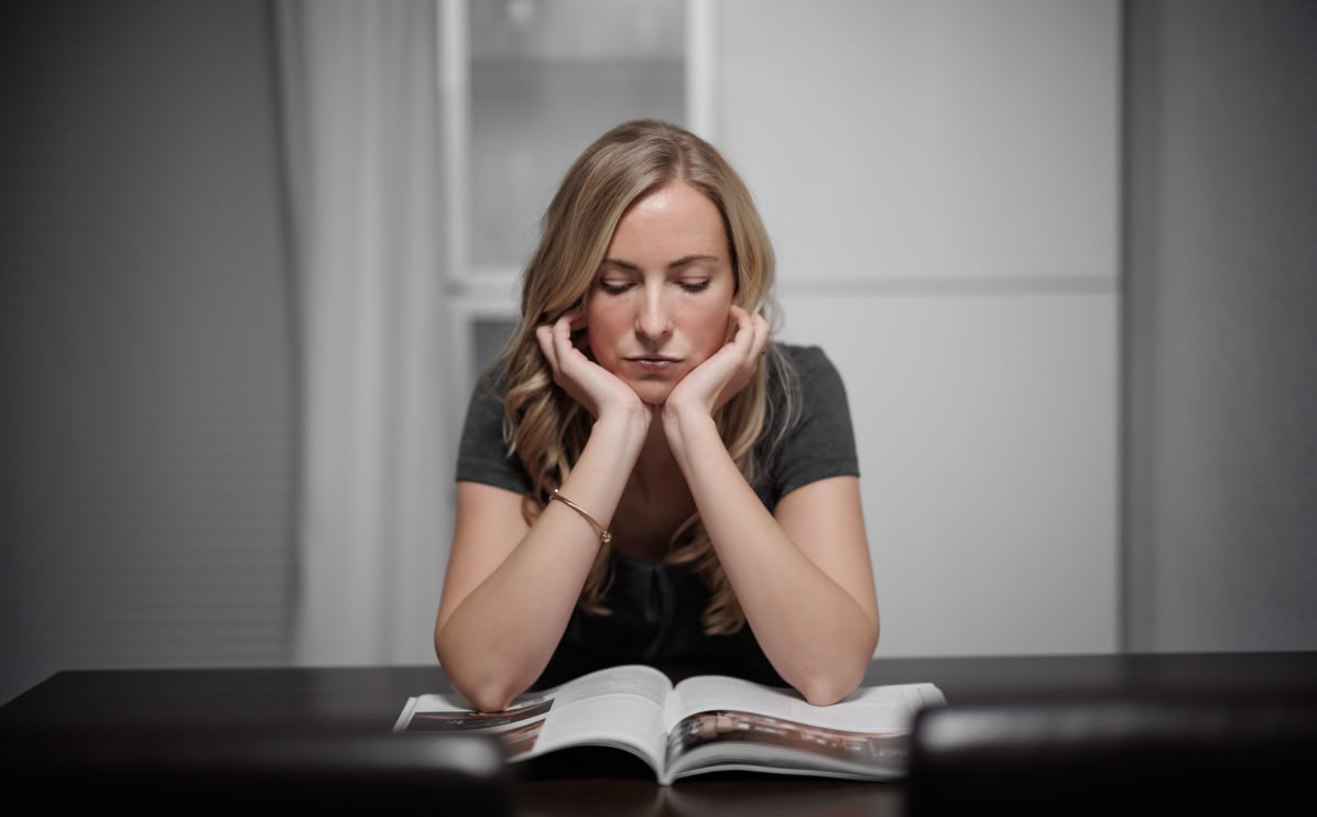 Frau mit langen Haaren sitzt an einem Tisch und stützt ihren Kopf auf die Hände, während sie auf ein geöffnetes Buch schaut.