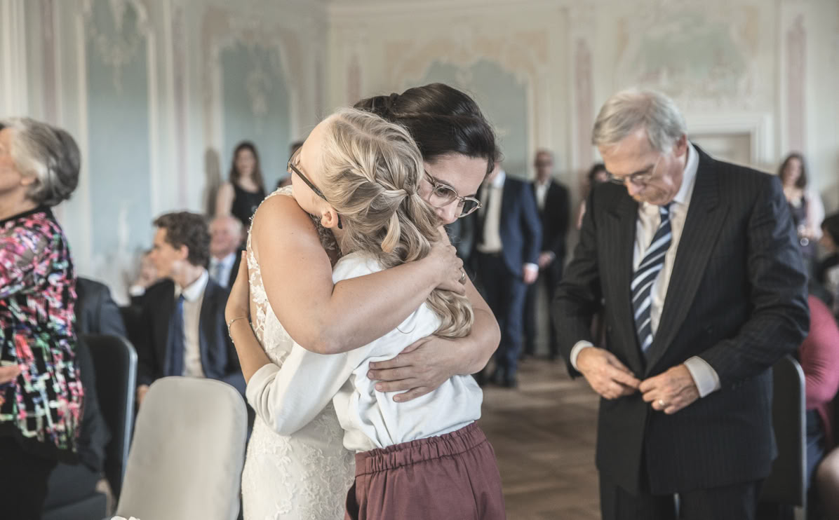 Zwei Personen umarmen sich in einem festlichen Raum, während andere Personen im Hintergrund sitzen und stehen.