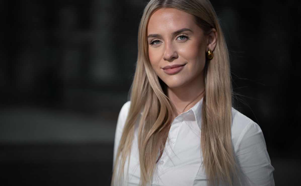 Eine Frau mit langen blonden Haaren trägt ein weißes Hemd und steht vor einem unscharfen Hintergrund.